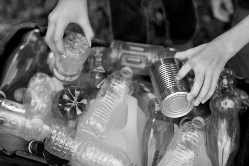 A close-up view of a person's hands sorting recyclable waste, with one hand holding a crushed metal can and the other reaching for a plastic bottle, both positioned over a collection of various discarded plastic bottles and cans. The plastic bottles are of different sizes, some with ridged textures and others smooth, with clear and lightly tinted surfaces. The metal can appears to be a standard aluminium drink can with a shiny finish. The background is blurred, emphasizing the focus on the waste items in the foreground. This scene likely takes place outdoors, related to waste sorting or rubbish collection, aligning with services offered by Office Clearance Tooting in south London, ensuring environmentally responsible disposal of waste and recyclable materials.