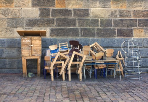 An outdoor scene featuring a variety of discarded furniture arranged against a weathered brick wall. The collection includes several wooden chairs with different finishes and styles, some stacked, others leaning or placed on the ground, along with a small wooden cabinet or cupboard with a slatted door situated on the left side. To the right, there are metal chairs and a small metal table, all showing signs of wear. The furniture is positioned on a cobbled pavement, suggesting an urban or premises clearance setting. The overall atmosphere is one of waste accumulation awaiting removal, aligning with rubbish removal services in Tooting or nearby areas. Office Clearance Tooting specializes in managing such clutter and unwanted items effectively, providing professional rubbish clearance support. The scene emphasizes the range of furniture items collected and the outdoor environment typical for rubbish disposal in a local borough context.