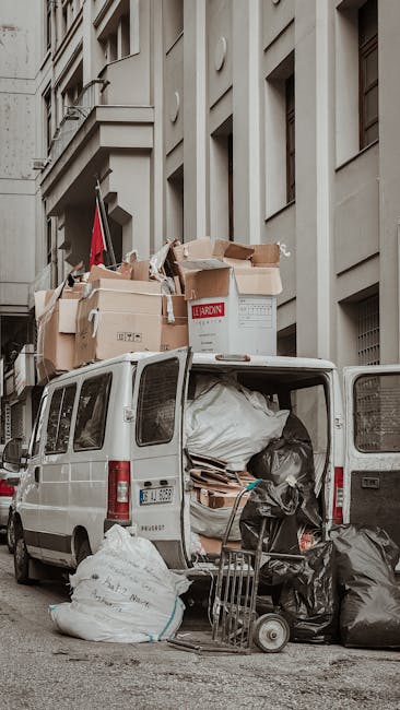 A worker wearing a white face mask and a light blue dress shirt is standing on the edge of a large, open-top waste collection truck, which is parked outdoors in an urban environment. The truck’s ind