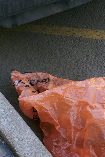 An orange plastic rubbish bag is lying on a textured asphalt surface in an outdoor setting, partially overlapping a concrete curb and adjacent to a building wall or pavement edge. The bag appears crum