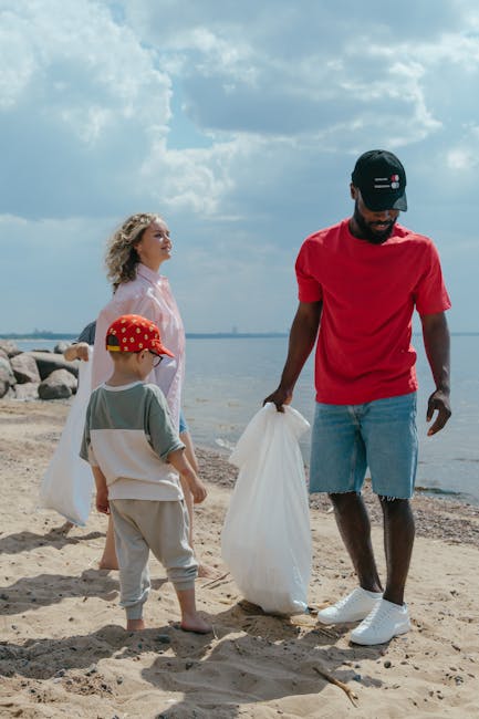 A person wearing a yellow and gray check shirt, gray pants, and bright green gloves is holding open a large black garbage bag, preparing for rubbish collection outdoors on a grassy area. The bag appea